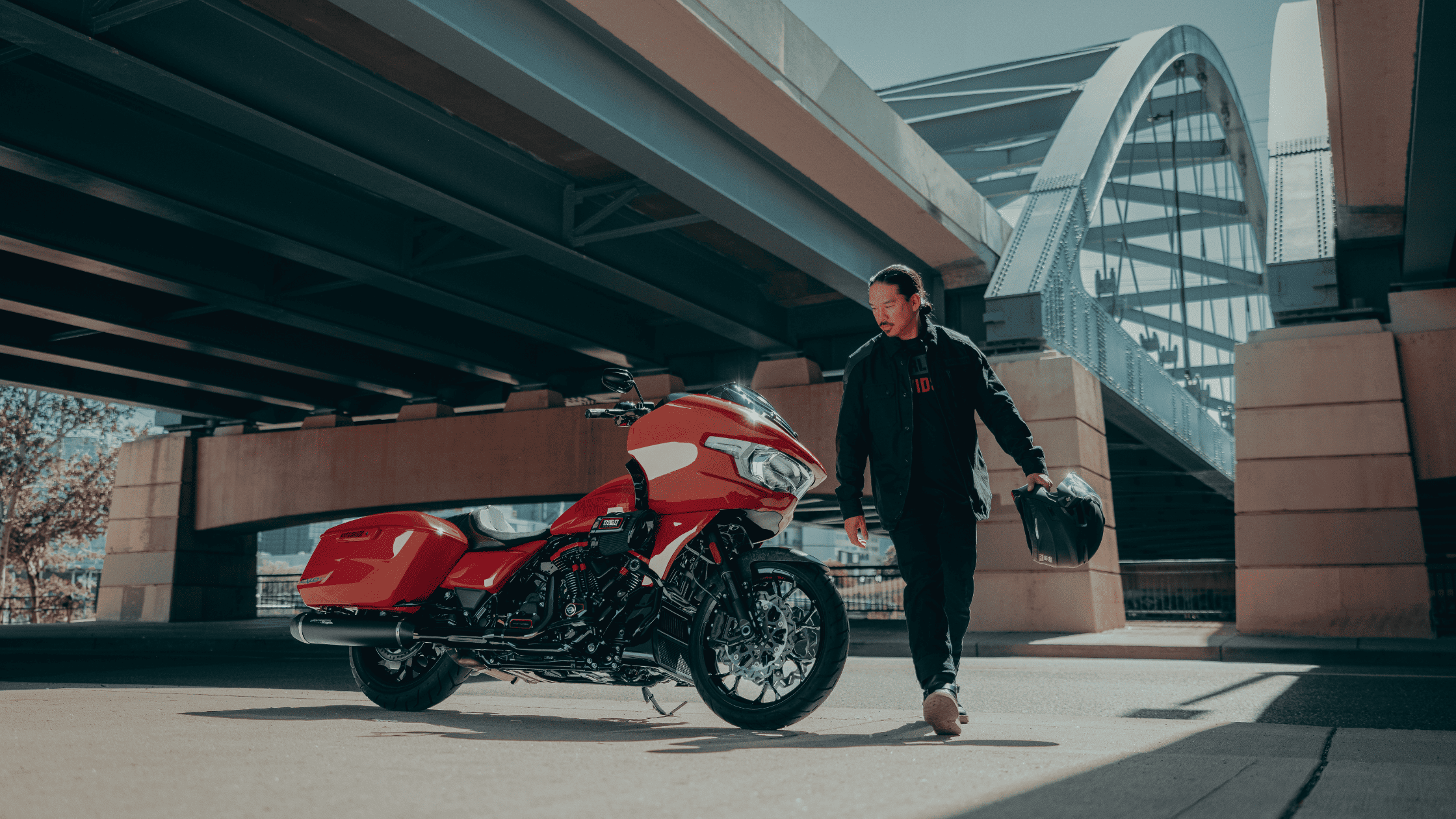 Rider walking toward a red 2025 Harley-Davidson CVO Road Glide ST parked under a modern city bridge.