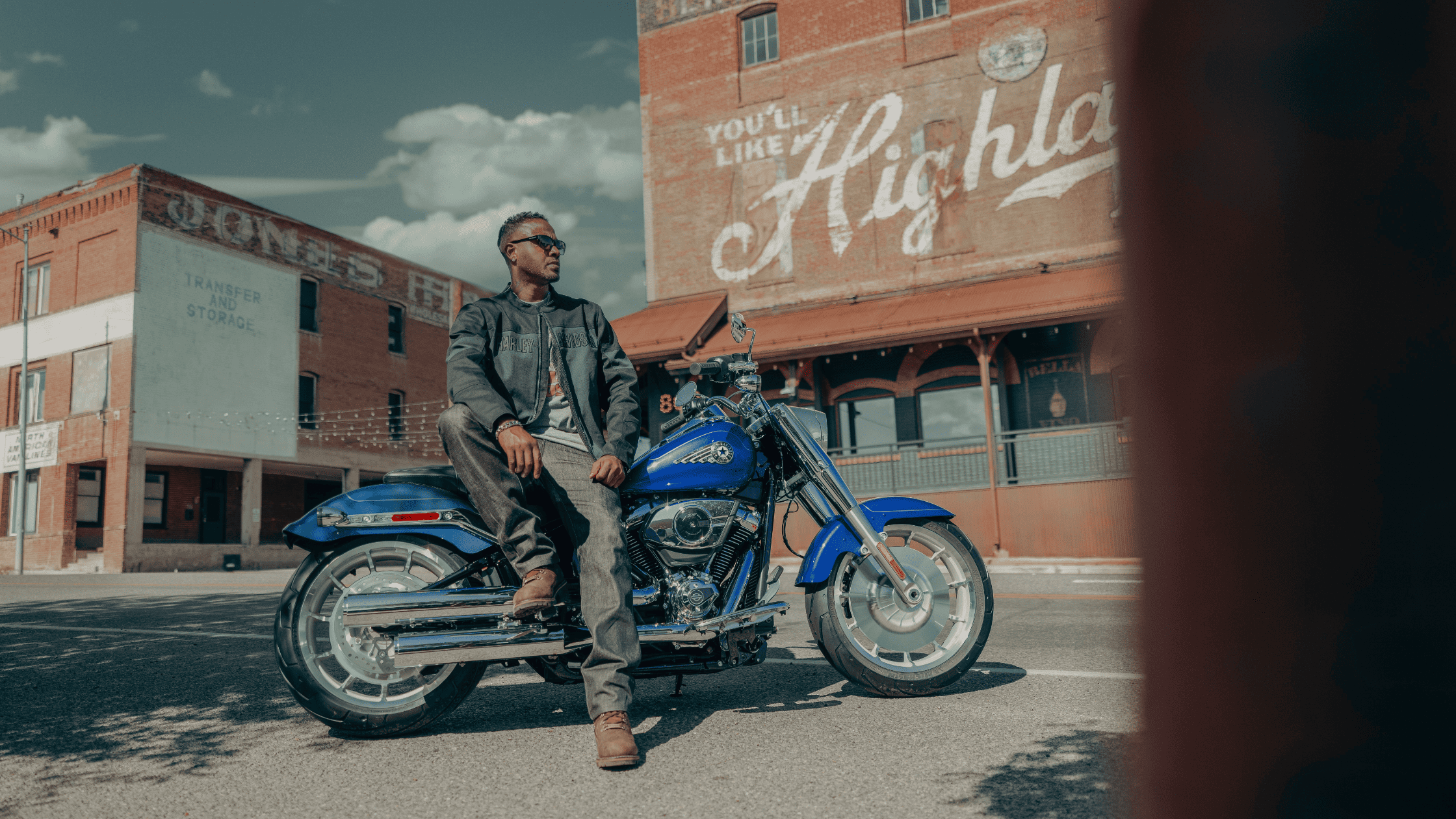 Man sitting on 2025 Harley-Davidson® Fat Boy® in front of vintage Highland mural on sunny street