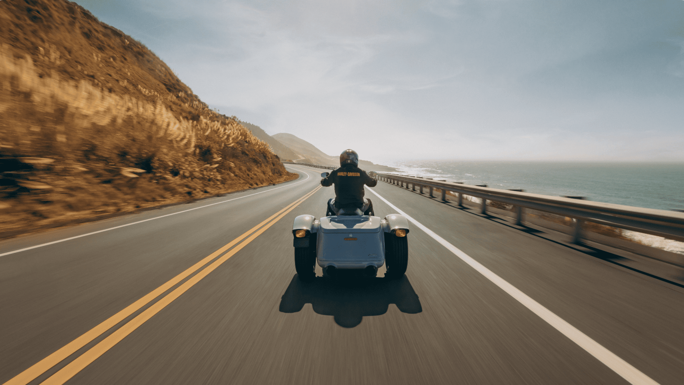 A rider drives the 2025 Harley-Davidson® Freewheeler® trike along a coastal highway with ocean views.