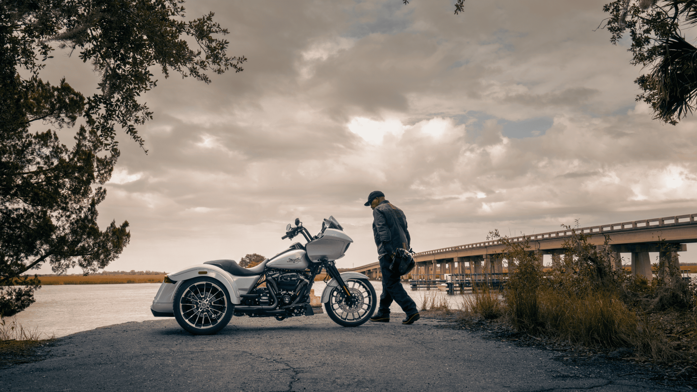 A person stands beside the 2025 Harley-Davidson® Road Glide® 3 trike near a bridge on an overcast day.